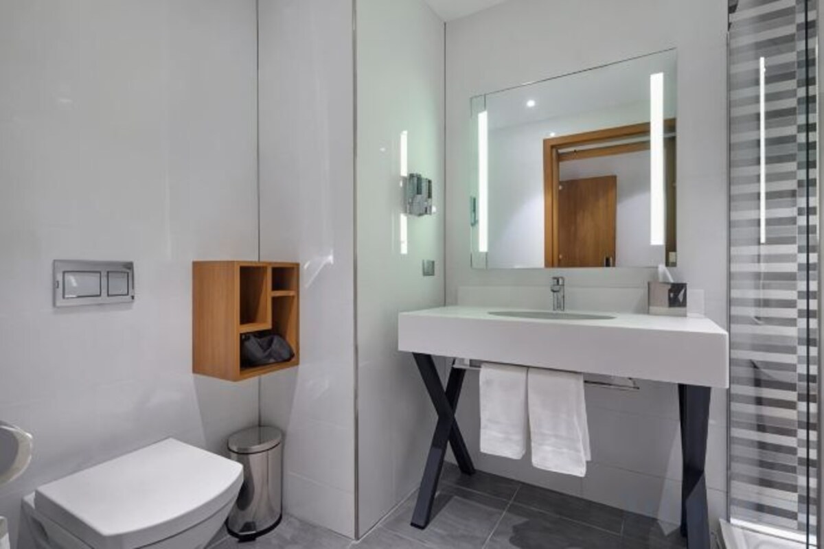 A modern bathroom with a white sink, a mirror, a towel rack, and a small wooden shelf.
