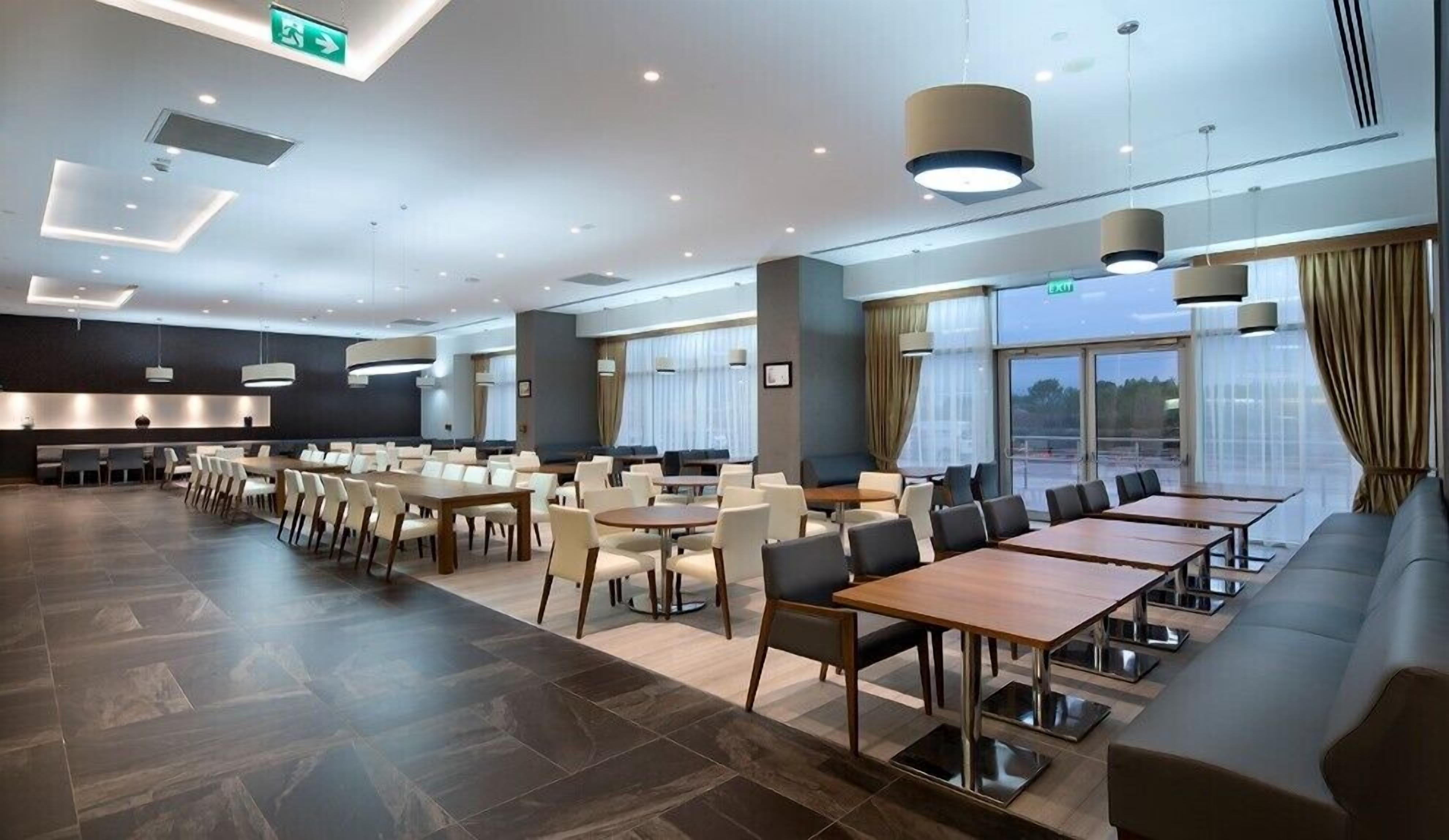 A modern hotel lobby with a long dining area, multiple seating options, and large windows offering views.