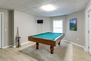 Una habitación con mesa de billar, televisor montado en la pared y ventana con persianas.