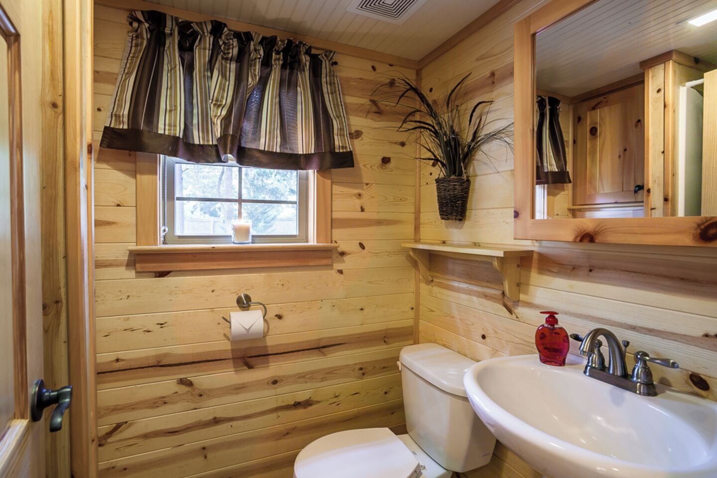 A wooden cabin bathroom with a toilet, sink, and window with curtains.
