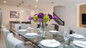 Un coin repas moderne avec une table en verre dressée pour quatre personnes, entourée de chaises blanches. Un vase contenant des fleurs violettes et jaunes est placé au centre.