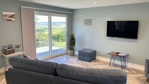 A modern living room with a grey sofa, a flat-screen TV, and a sliding glass door leading to a balcony with a view of greenery. - Secure, Detached bungalow overlooking Weston-super-Mare, the Severn and Wales (Hutton)