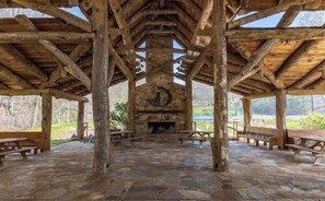 A covered pavilion with a stone floor, wooden benches, and a central fireplace. - 2 mins to Ski! Hot Tub+Fire Pit, Fireplace, Grill! (Mars Hill)