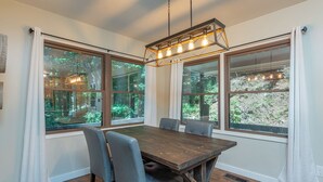 A dining area with a wooden table, six chairs, and large windows offering a view of greenery. - Come Ski Hatley Pointe! Hot Tub+Fire Pit, Fireplace,WiFi, Pet Friendly!  (Mars Hill)