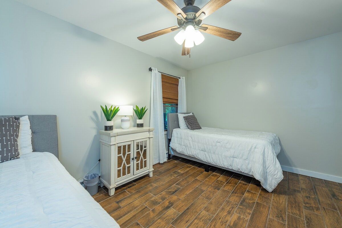 A bedroom with two beds, a ceiling fan, a cabinet with plants, and a window with curtains.