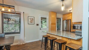 A modern kitchen with wooden cabinets, a stainless steel refrigerator, and a granite countertop. There are three wooden stools and a dining table with chairs. - Come Ski Hatley Pointe! Hot Tub+Fire Pit, Fireplace,WiFi, Pet Friendly!  (Mars Hill)