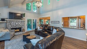 A spacious living room with a stone fireplace, leather sofas, a large flat-screen TV, and a dining area with natural light from large windows. - Come Ski Hatley Pointe! Hot Tub+Fire Pit, Fireplace,WiFi, Pet Friendly!  (Mars Hill)