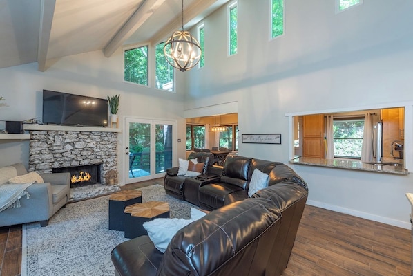 A spacious living room with a stone fireplace, leather sofas, a flat-screen TV, and a large window overlooking a deck. - 2 mins to Ski! Hot Tub+Fire Pit, Fireplace, Grill! (Mars Hill)