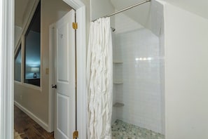 A bathroom with white tiled walls, a shower with a white curtain, and a visible door leading to another room. - 2 mins to Ski! Hot Tub+Fire Pit, Fireplace, Grill! (Mars Hill)
