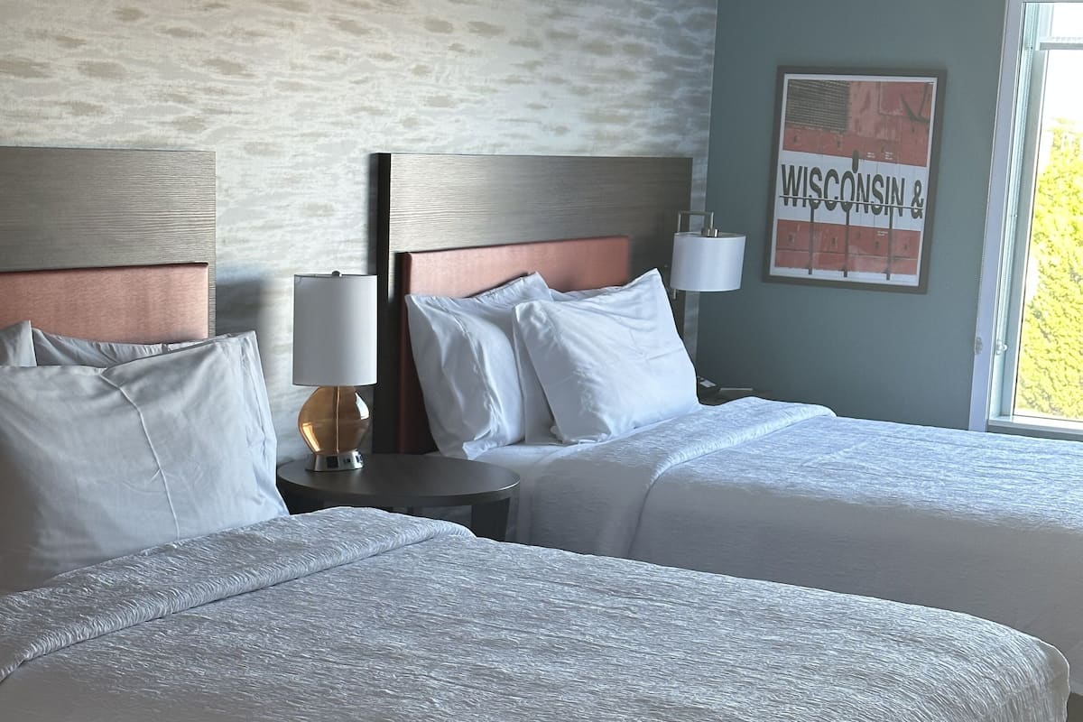 A hotel room with two beds, a nightstand with a lamp, a framed picture on the wall, and a window with a view.