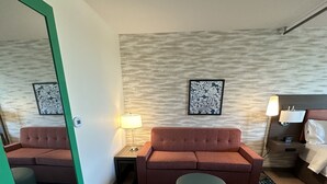 A hotel room with a brown sofa, a blue ottoman, a patterned rug, and two wall-mounted lamps. - Home2 Suites By Hilton Racine (Sturtevant)