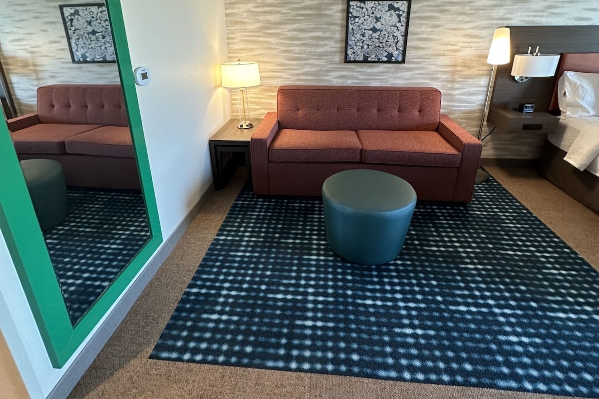 A hotel room with a brown sofa, a blue ottoman, a patterned rug, and two wall-mounted lamps.