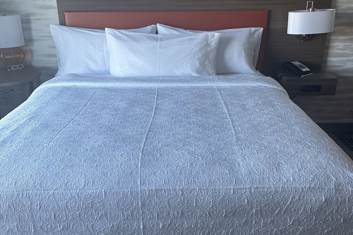 A neatly made bed with white linens, a wooden headboard, and a nightstand with a lamp and phone.