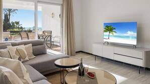 A modern living room with a sofa, a glass coffee table, a TV displaying a beach scene, and a balcony with a view of the ocean.