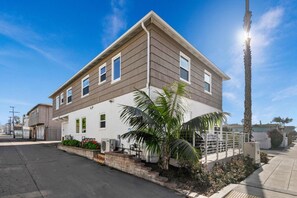 A two-story residential building with a flat roof, a front porch, and a small garden area with palm trees and flowers.