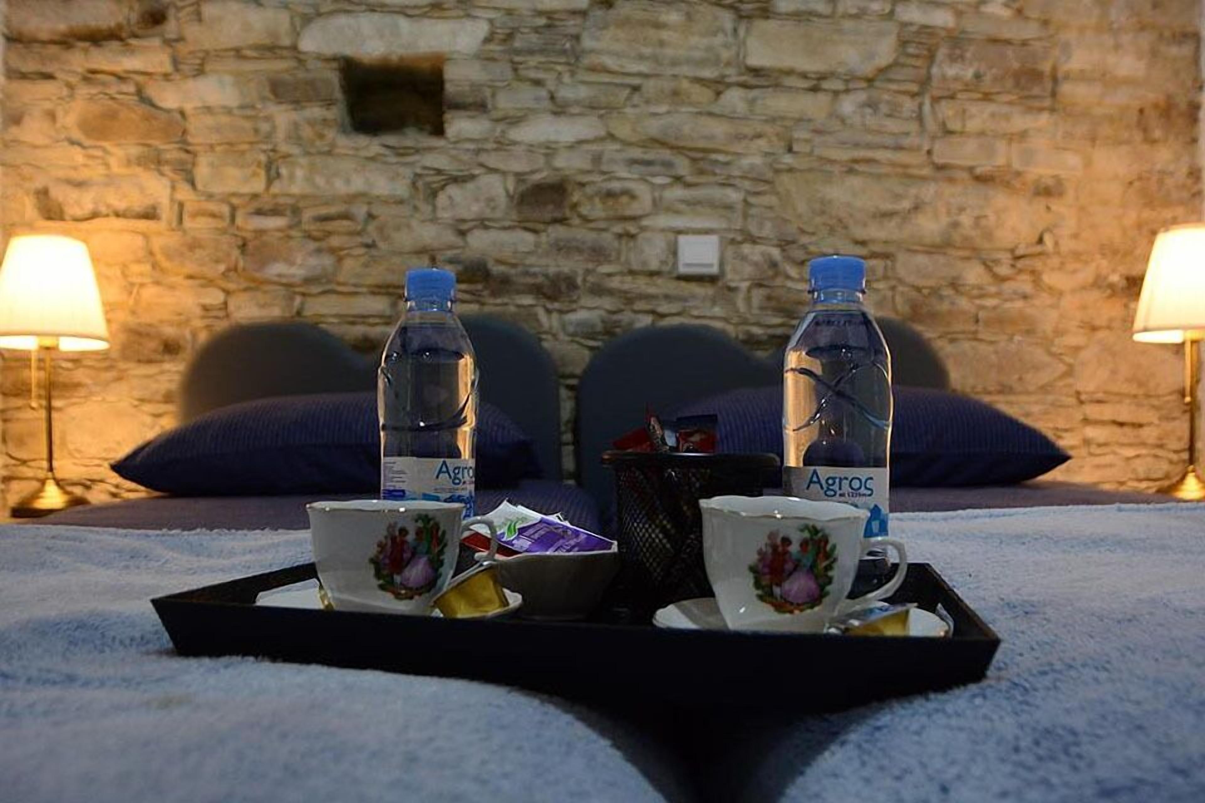 A cozy room with a stone wall, two lamps, a tray with cups and a bottle, and a bed with blue pillows.