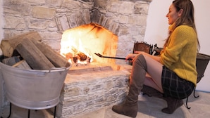 A person sitting by a stone fireplace with logs, a metal bucket, and a poker. - Lazaros Stonehouse (Pano Lefkara)