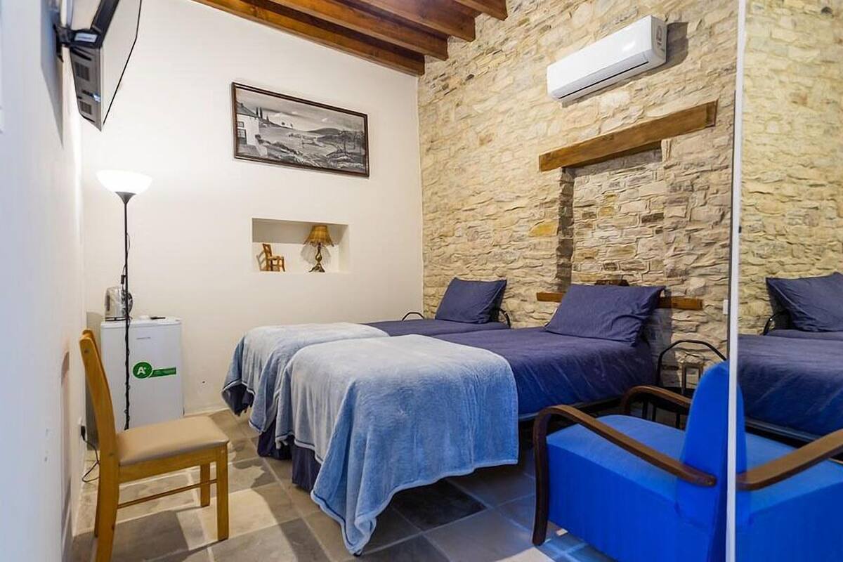 A room with two beds, a chair, a desk with a lamp, a small table, and a stone wall.
