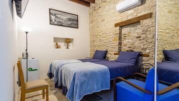 A room with two beds, a chair, a desk with a lamp, a small table, and a stone wall.