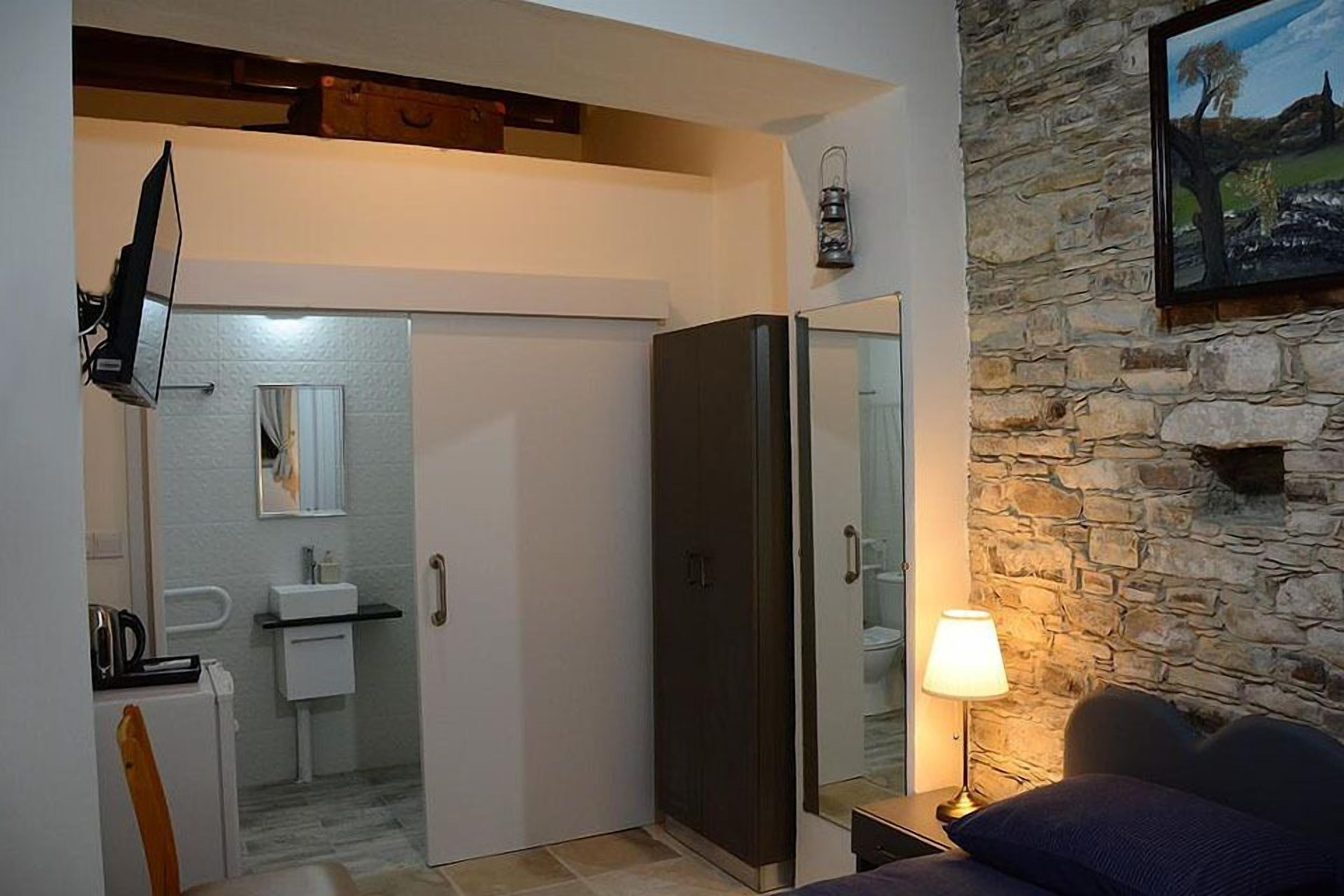 Un dormitorio con cama, mesita de noche con lámpara, armario, espejo, televisor montado en la pared y pared de piedra.