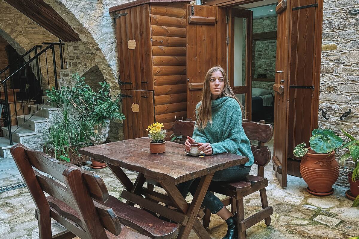 A rustic wooden table and chairs set in a stone courtyard with a wooden cabin and a stone wall in the background.