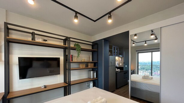 Una habitación de hotel moderna con cama, televisión de pantalla plana, estanterías integradas, zona de cocina y balcón con vistas a la ciudad.
