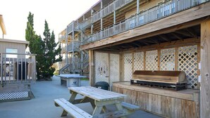 Een houten terras met een picknicktafel en barbecue, omgeven door een houten hek en een gebouw met balkons.