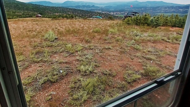 A view from inside a vehicle, looking out through a window at a grassy field with distant mountains and a few buildings.