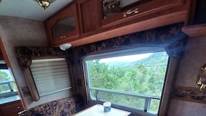 A compact RV interior with a dining area, a small table, and a window offering a view of greenery.