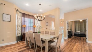 Un coin repas avec une table et des chaises en bois, un lustre et une vue sur une chambre à coucher par une porte ouverte.