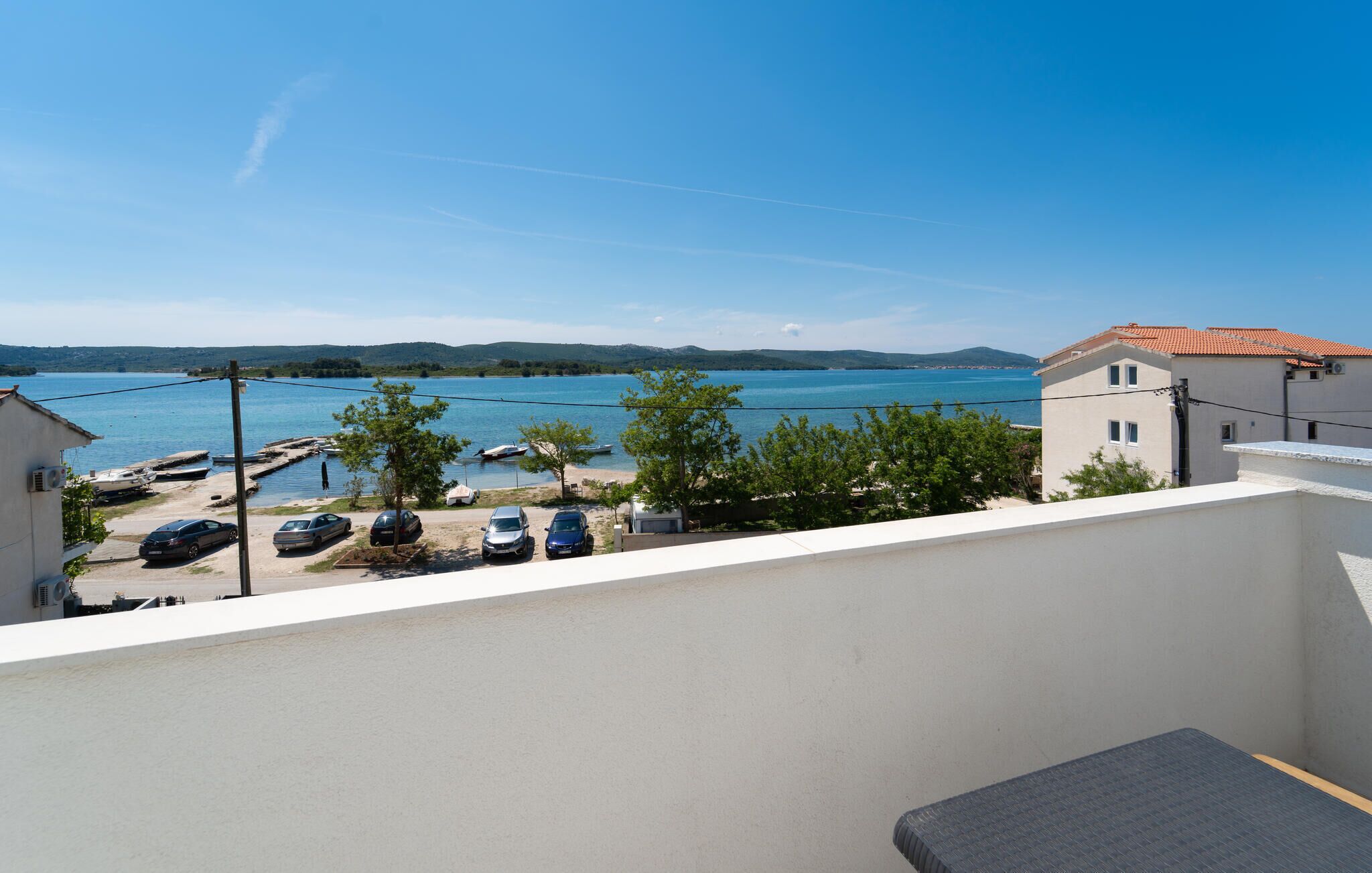 A balcony with a view of a lake, parked cars, and a building with a tiled roof.