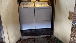 Une buanderie équipée de deux machines à laver et deux sèche-linge superposés, d’une étagère contenant des produits de lessive et d’une porte en bois.