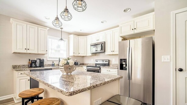 A modern kitchen with white cabinetry, granite countertops, stainless steel appliances, and a central island with stools. - The Clemson Cottage - 3.6 mi. to Memorial Stadium (Seneca)