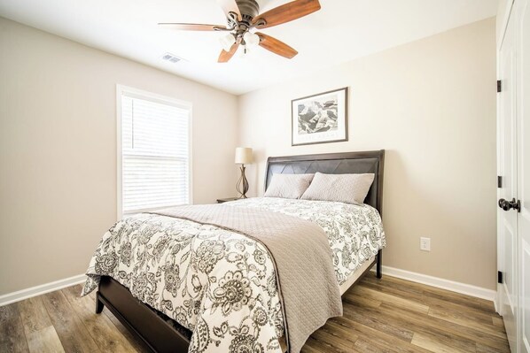 A bedroom with a bed, a ceiling fan, a window with blinds, a bedside table with a lamp, and a framed picture on the wall. - The Clemson Cottage - 3.6 mi. to Memorial Stadium (Seneca)