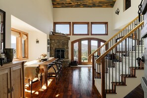 A spacious kitchen with a stone fireplace, wooden flooring, and a view through large windows.