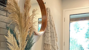 A modern interior with a glass vase of pampas grass, a wooden-framed mirror, a white chair, and a stone fireplace.