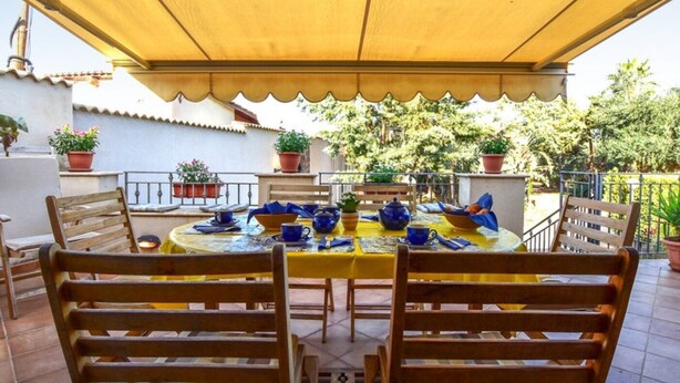 A covered outdoor dining area with a table set for four, surrounded by wooden chairs, and potted plants on the terrace. - Baglio Cuttaia - Apartment for families. (Carini)