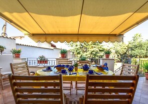 An outdoor patio with a round table, wooden chairs, and a yellow tablecloth.