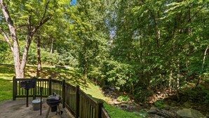 A deck with a grill and a table, surrounded by a wooden fence and lush greenery. - Those Who Wander Are Not Always Lost – Cozy Mountain Stream Condo with Fireplace (Gatlinburg)