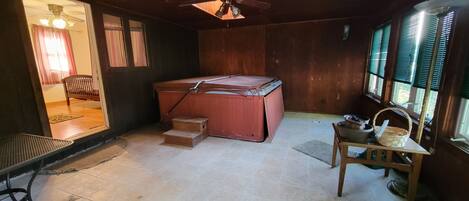A room with a wooden ceiling, a ceiling fan, a hot tub, a small wooden table with a basket, and a window with blinds.