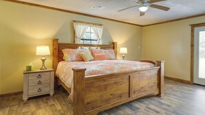 Un dormitorio con una cama de madera, dos mesitas de noche, una mesita auxiliar, un ventilador de techo y una ventana con cortinas.