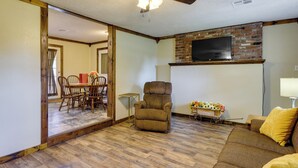 Una sala de estar con piso de madera, un sofá, un sillón reclinable, una mesa de comedor con sillas y un televisor montado en una pared de ladrillo.