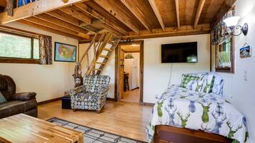 A cozy room with a wooden ceiling, a bed with a deer print duvet, a brown leather sofa, a patterned armchair, and a wooden coffee table.