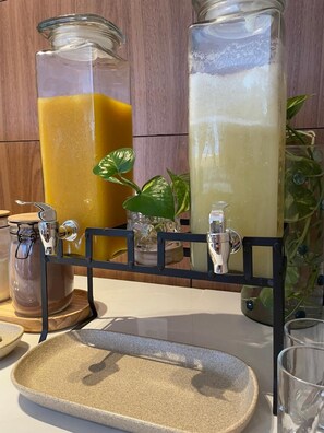 A kitchen counter with two glass jars containing orange and white liquids, a metal dispenser, and a ceramic tray. - Blue Hotel (João Pessoa)