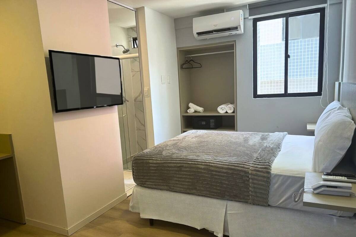 A modern bedroom with a bed, a TV mounted on the wall, a wardrobe, and an air conditioning unit.