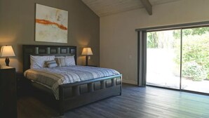 A bedroom with a large bed, two bedside lamps, a ceiling with exposed beams, and a large sliding glass door leading to an outdoor area. - Modern Mission Hills Country Club Townhome (Rancho Mirage)