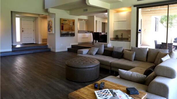 A spacious living area with a large sectional sofa, a round ottoman, and a coffee table. There is a kitchen in the background with white cabinets and a dining area. - Modern Mission Hills Country Club Townhome (Rancho Mirage)