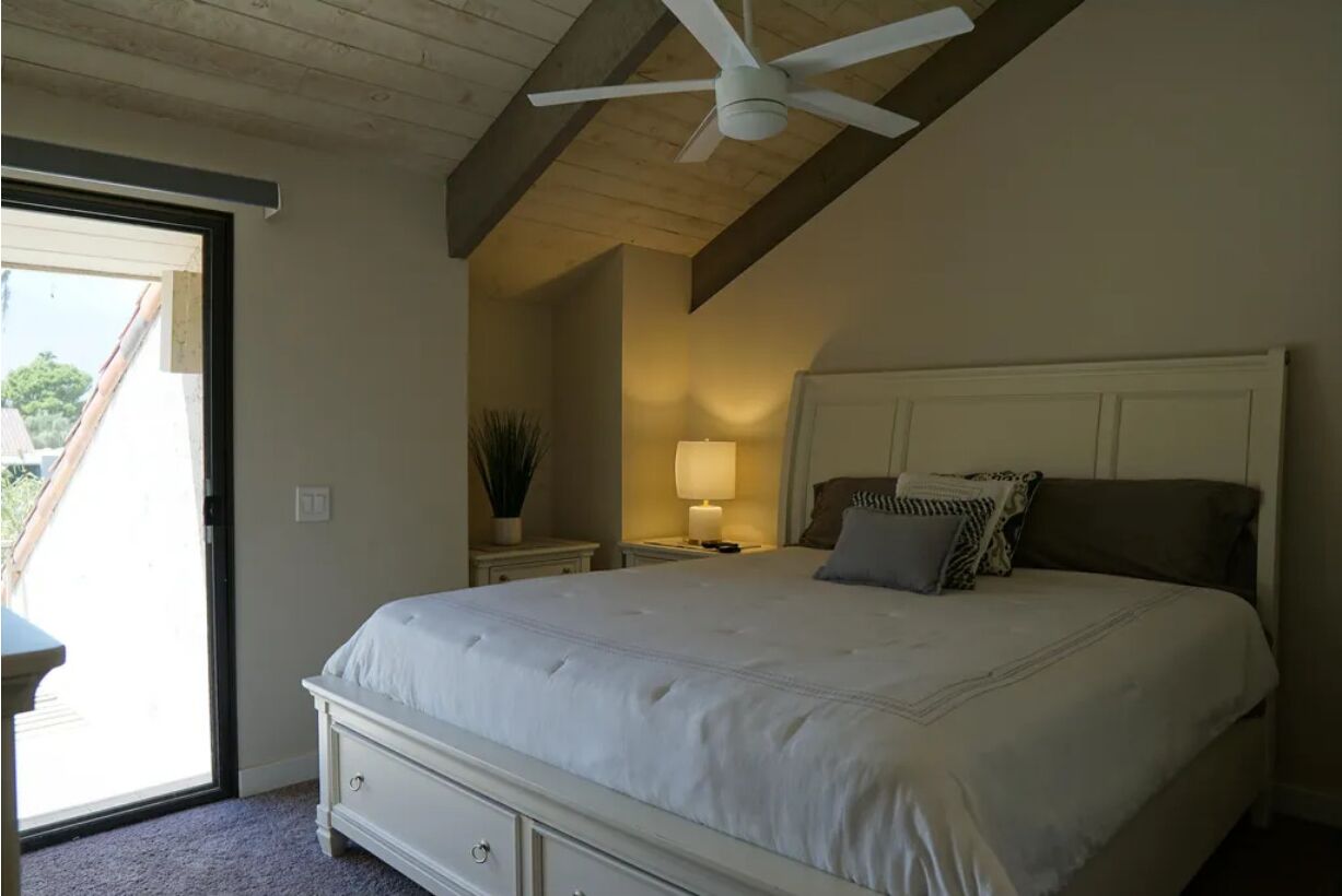 Een slaapkamer met een bed, een plafondventilator, een nachtkastje met een lamp, en uitzicht op de omgeving.