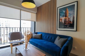 A modern living room with a blue sofa, a grey chair, and a wooden coffee table. There is a framed cityscape painting on the wall and a large window with blinds. - Wynwood Residencial in Miraflores (Lima)