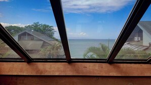 Blick durch ein Fenster mit Holzrahmen auf einen Strand mit Häusern und Palmen.